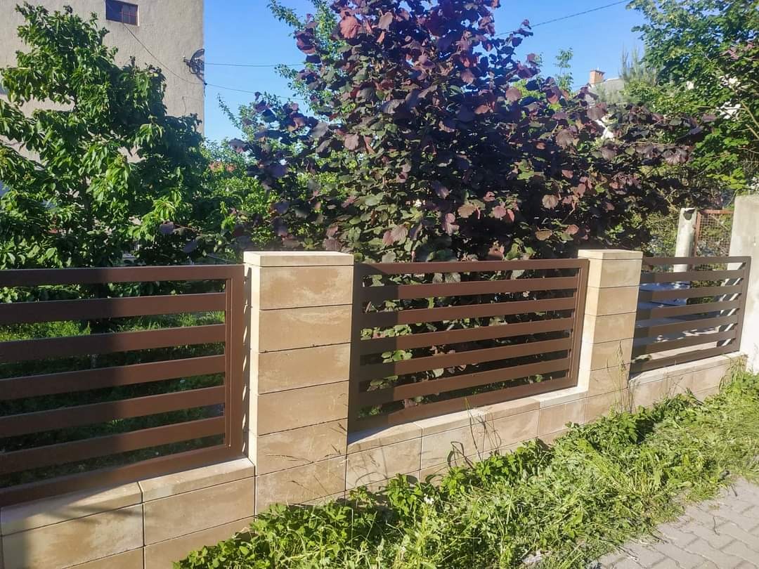 Nowoczesne ogrodzenie panelowe w Wiesiółce z poziomymi, brązowymi przęsłami, osadzone na murku z beżowych bloczków betonowych, na tle zieleni i drzew.