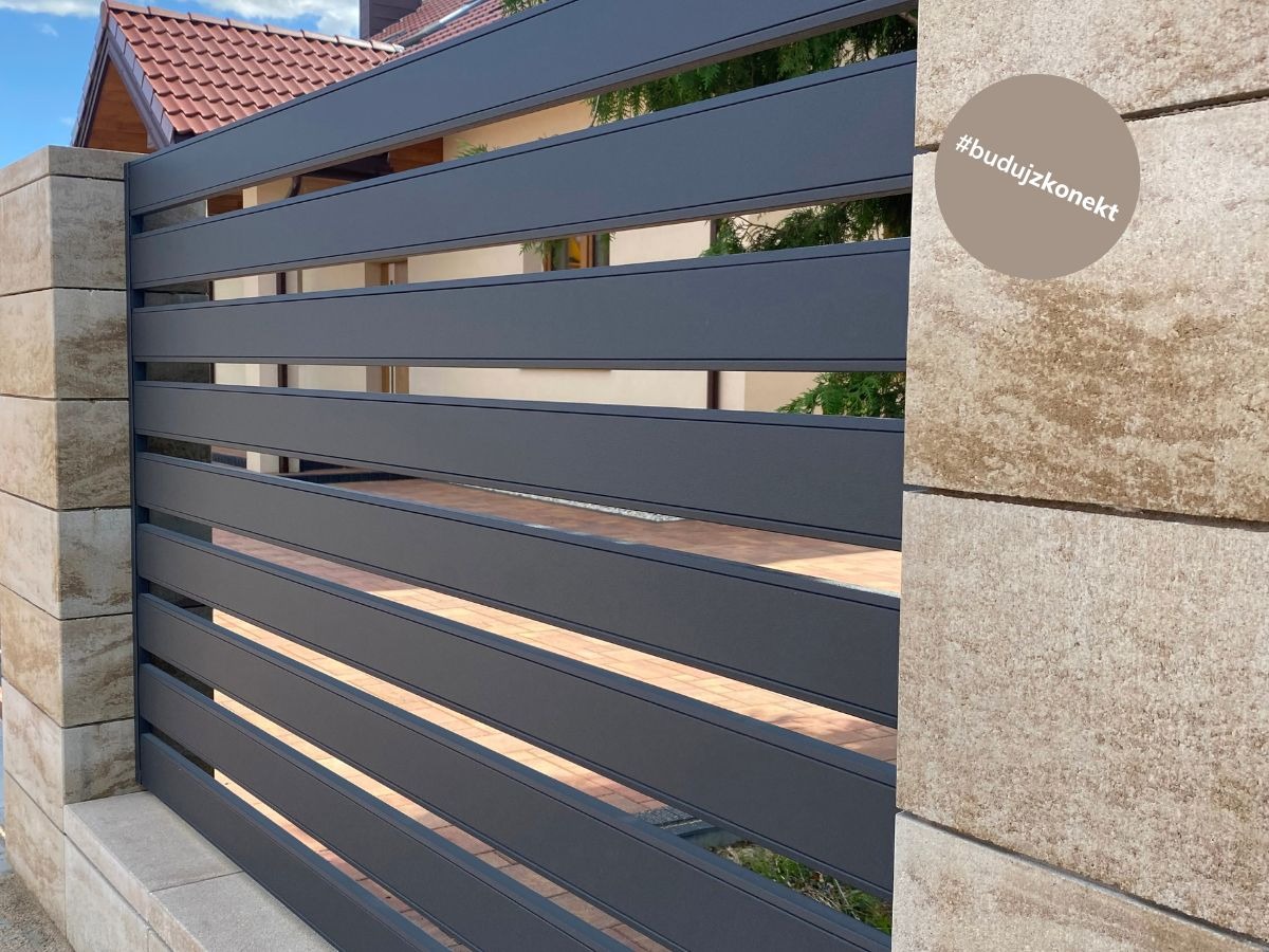 Nowoczesne ogrodzenie z poziomych, grafitowych paneli metalowych, osadzone na słupkach z jasnego kamienia z widocznym hashtagiem #budujzkonekt.