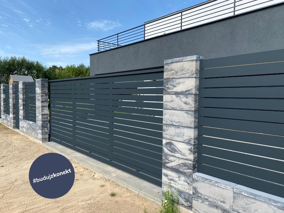 Nowoczesne, poziome przęsła w kolorze grafitowym, z elementami betonowymi o wzorze marmurkowym, tworzące ogrodzenie posesji na tle błękitnego nieba.
