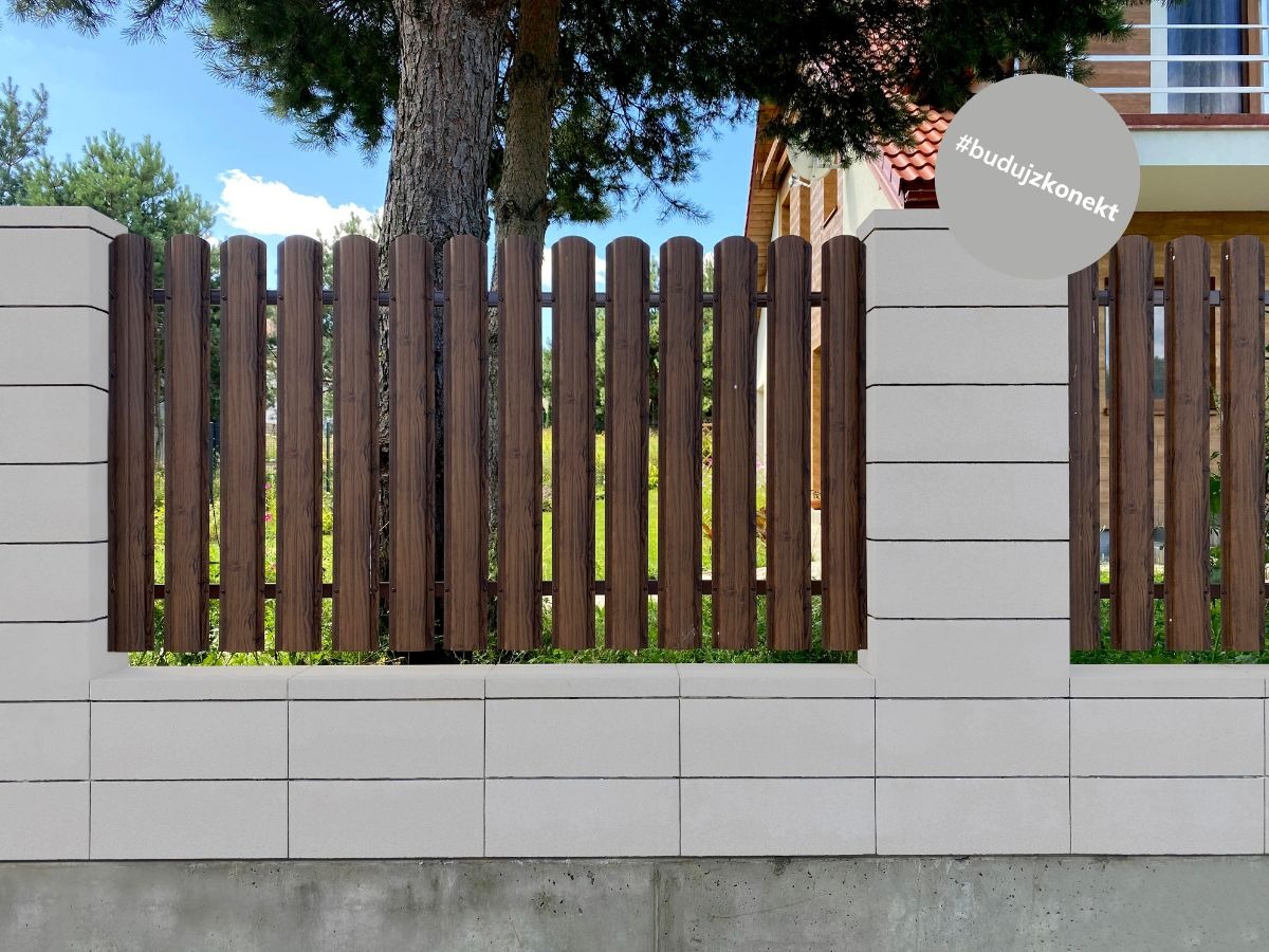 Fragment ogrodzenia z drewnianymi sztachetami i betonowymi słupkami, widok na zieleń i dom w tle, na słupku naklejka z hashtagiem #budujzkonekt.