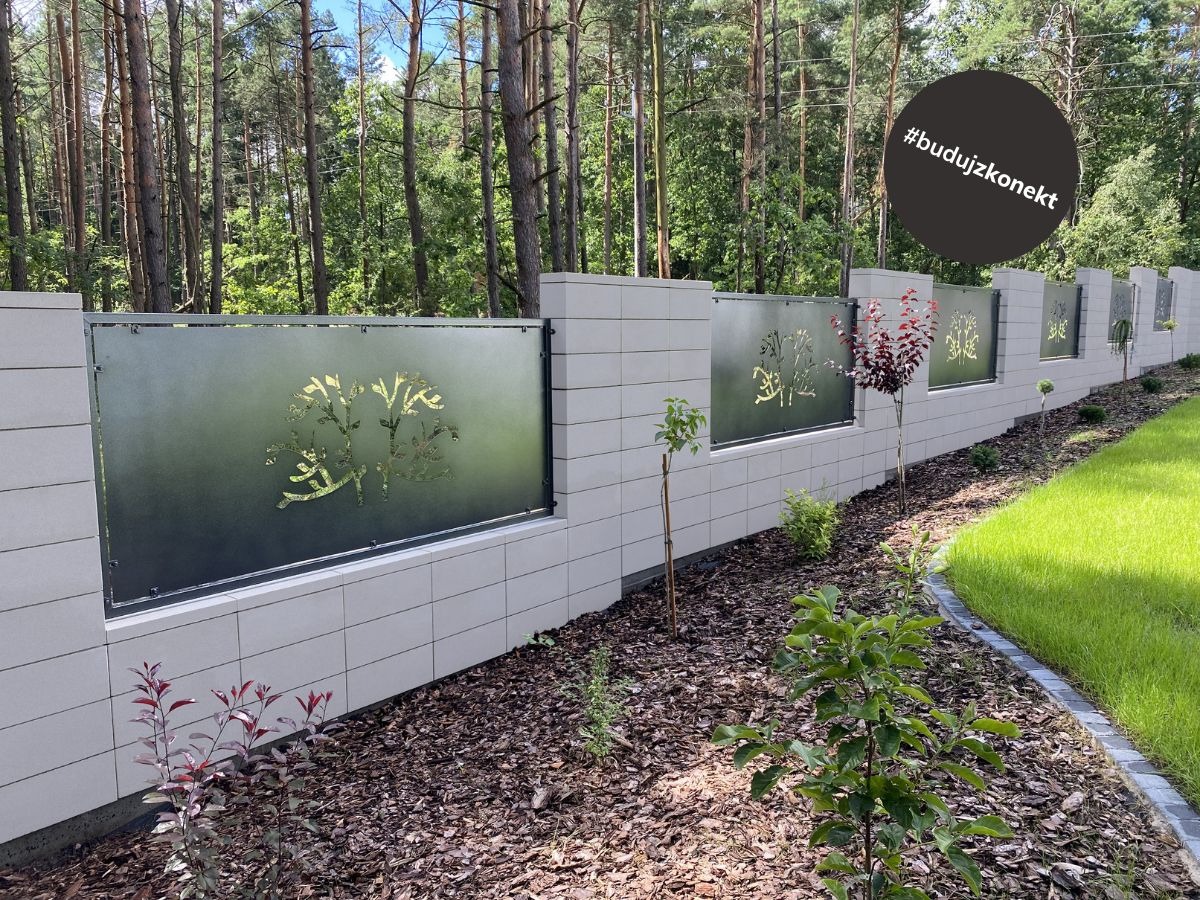 Nowoczesne ogrodzenie z betonowych bloczków z panelami z motywem drzewa, widok z zewnątrz w słoneczny dzień.