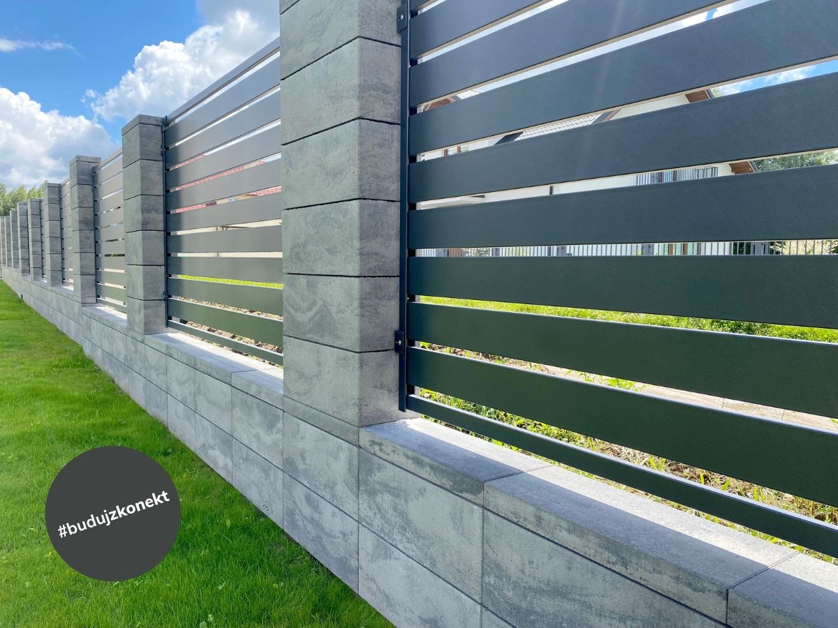 Nowoczesne ogrodzenie z poziomych, ciemnych paneli metalowych i szarych, betonowych słupków, widoczne z poziomu trawnika w słoneczny dzień.