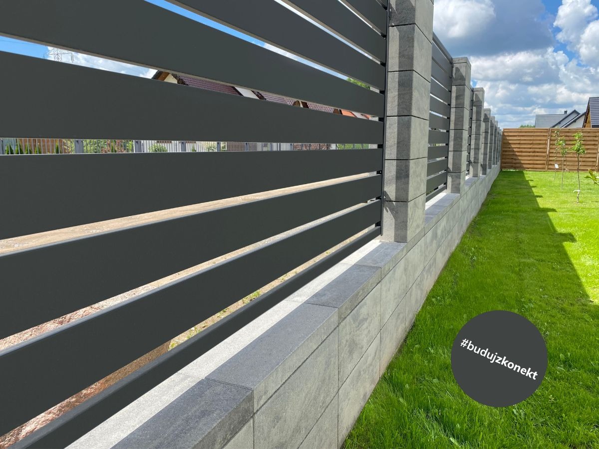 Nowoczesne ogrodzenie posesji z poziomymi, grafitowymi panelami i betonową podmurówką, widok z poziomu trawnika w słoneczny dzień.