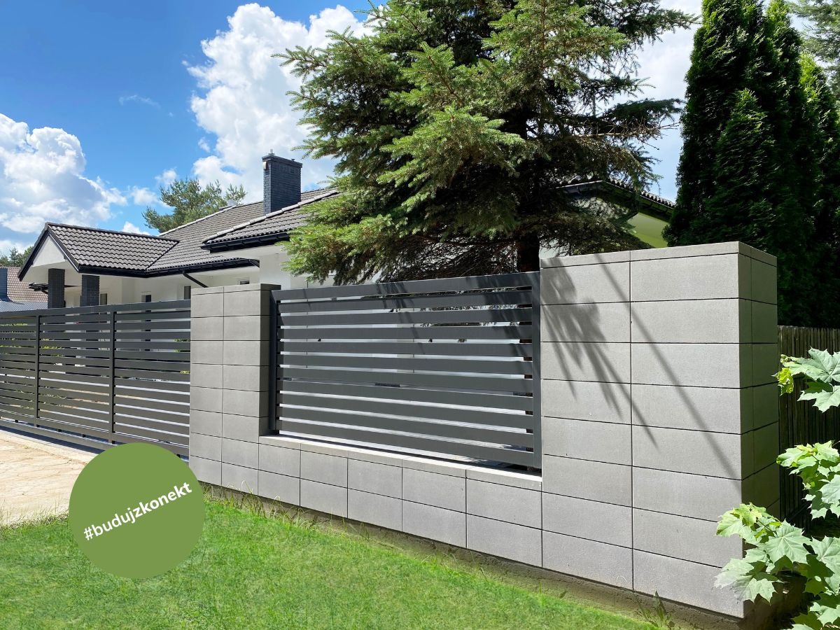 Nowoczesne ogrodzenie z betonowych bloczków i poziomych metalowych przęseł, z widocznym fragmentem domu z ciemną dachówką w tle, na tle błękitnego nieba z chmurami.