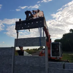 Montaż prefabrykowanych elementów betonowych ogrodzenia przy użyciu koparki, pracownik w rękawicach pomaga w ustawieniu elementu, w tle błękitne niebo z chmurami.