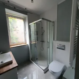 Nowoczesna łazienka po remoncie: szara glazura podłogowa, kabina prysznicowa z przezroczystego szkła, podwieszana toaleta, umywalka nablatowa na drewnianym blacie, okno z widokiem na zieleń.