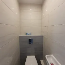 Wnętrze wykończonej łazienki z beżowo-szarymi płytkami, wiszącą toaletą z chromowanym przyciskiem spłukiwania, wąską umywalką z czerwonym mydłem w płynie i kratką wentylacyjną pod sufitem.