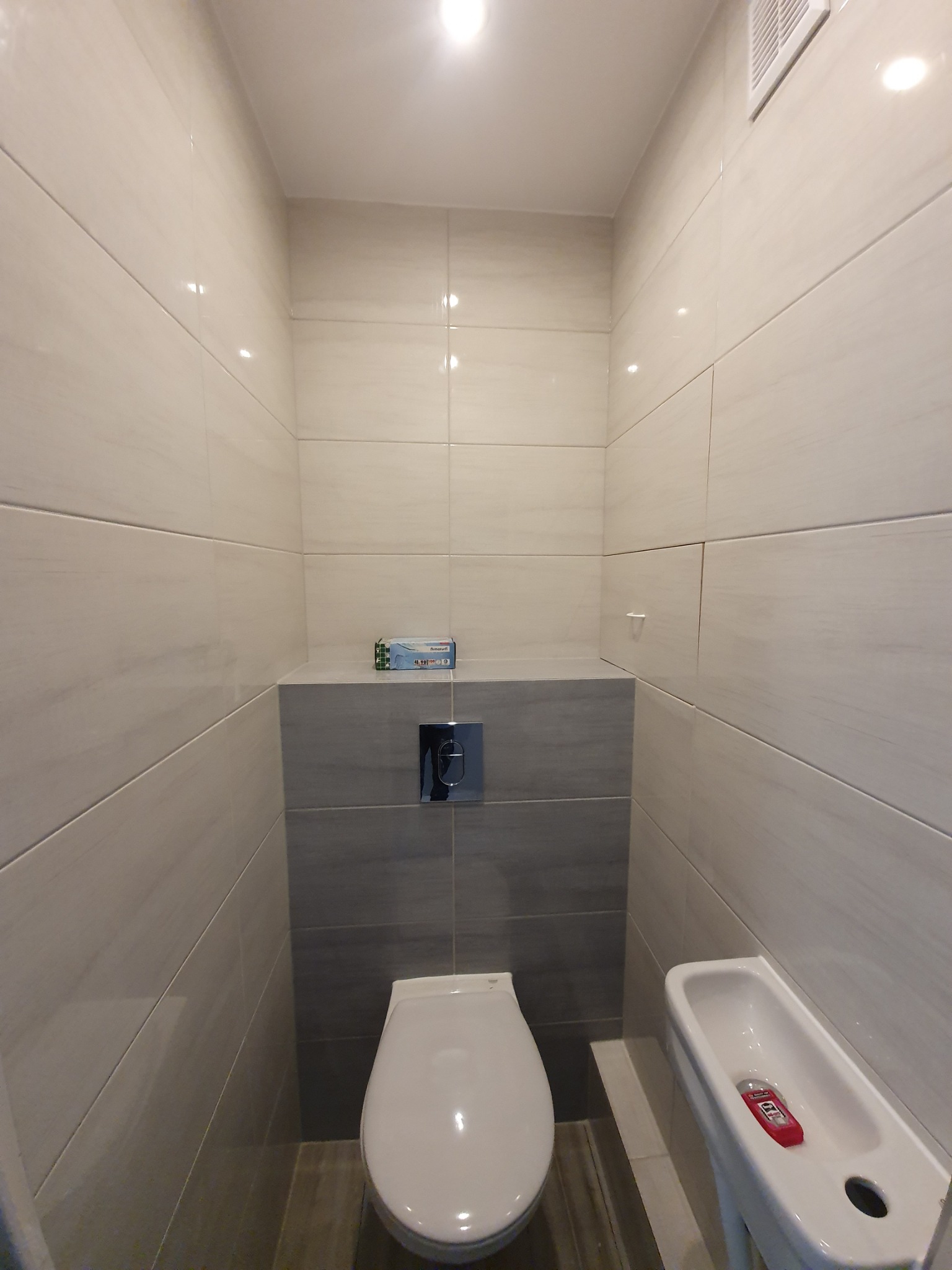 Wnętrze wykończonej łazienki z beżowo-szarymi płytkami, wiszącą toaletą z chromowanym przyciskiem spłukiwania, wąską umywalką z czerwonym mydłem w płynie i kratką wentylacyjną pod sufitem.