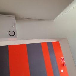 SIROSYSTEM - Biała skrzynka z okrągłym logo na ścianie, nad wejściem z pomarańczowo-szarymi pionowymi pasami i symbolem ostrzegawczym z czerwoną błyskawicą.