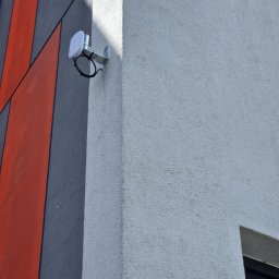 SIROSYSTEM - Biała antena Wi-Fi zamontowana na metalowym uchwycie na rogu budynku z szarą i pomarańczową dekoracją.