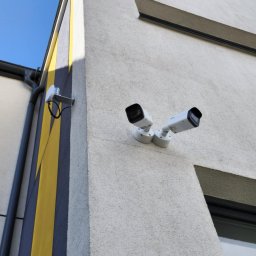 SIROSYSTEM - Dwie białe kamery monitoringu na elewacji budynku z żółto-szarym pasem i anteną Wi-Fi, skierowane w przeciwne strony.
