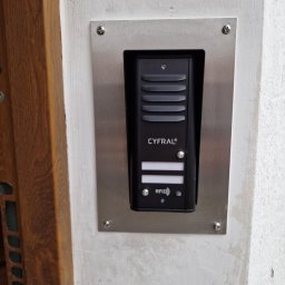 SIROSYSTEM - Czarny wideofon marki Cyfral zamontowany na srebrnej, metalowej płycie na pomalowanej, białej ścianie obok drewnianej framugi drzwi.