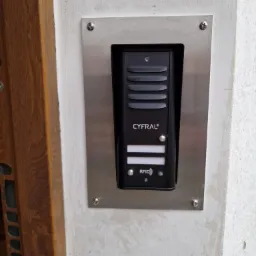 Czarny wideofon marki Cyfral zamontowany na srebrnej, metalowej płycie na pomalowanej, białej ścianie obok drewnianej framugi drzwi.