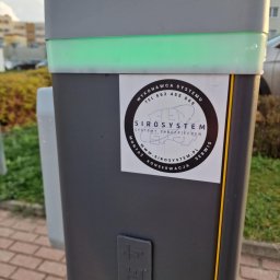 SIROSYSTEM - Szary słupek z zielonym podświetleniem i naklejką firmy SIROSYSTEM, zajmującej się systemami zabezpieczeń, widoczny na zewnątrz budynku.