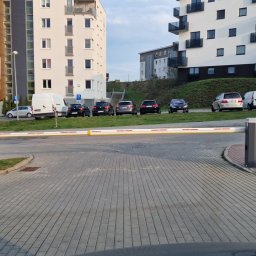 SIROSYSTEM - Szlaban automatyczny z żółto-białymi pasami na tle nowoczesnego osiedla mieszkaniowego, zaparkowane samochody, kostka brukowa.