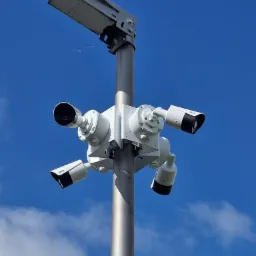 Kompleksowy system monitoringu: cztery kamery CCTV zamontowane na metalowym słupie oświetleniowym, na tle błękitnego nieba z chmurami. Widok z dołu.