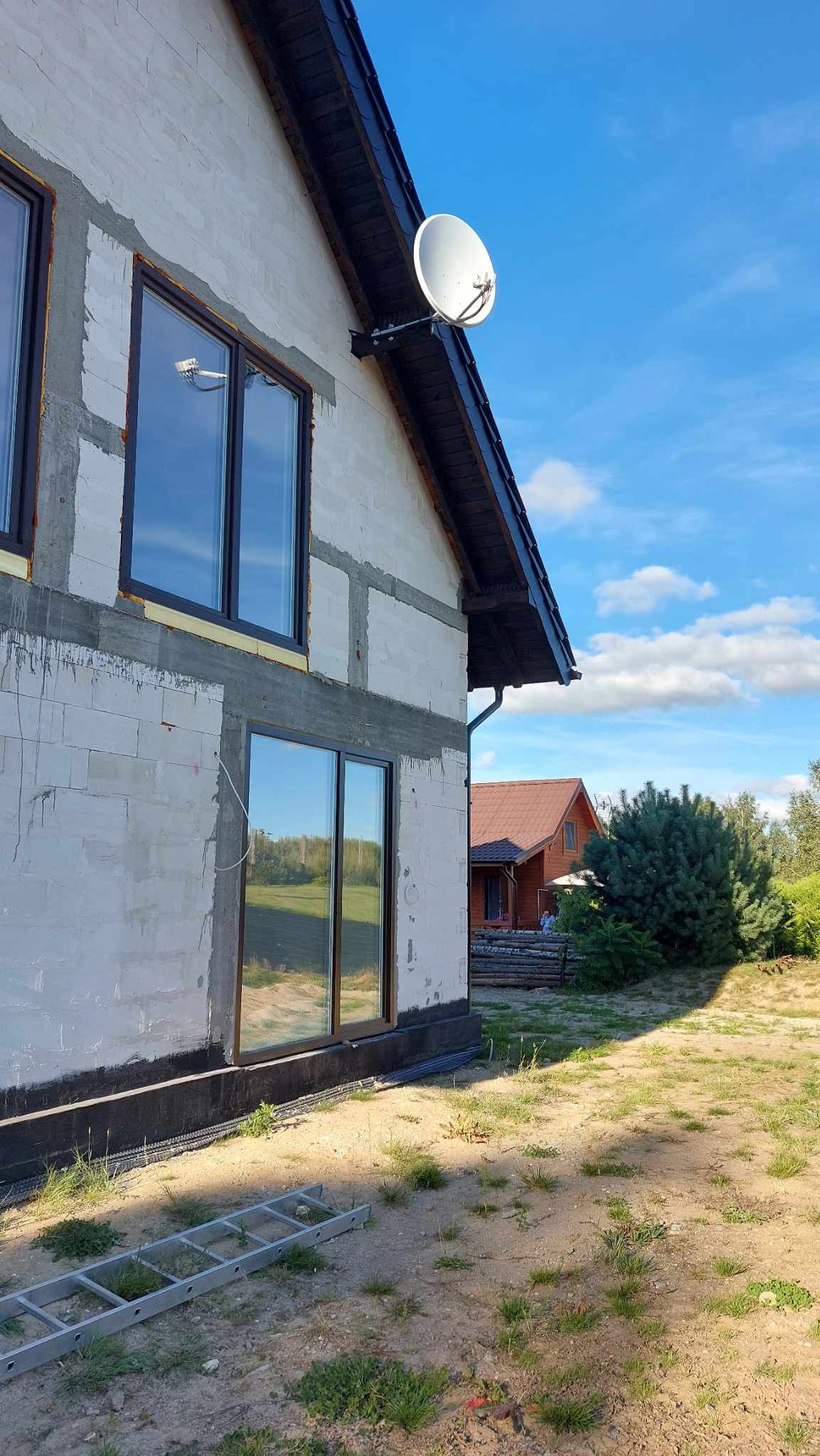 Antena satelitarna zamontowana na ścianie surowego domu z oknami z czarnymi ramami, widoczna drabina oparta o ścianę.