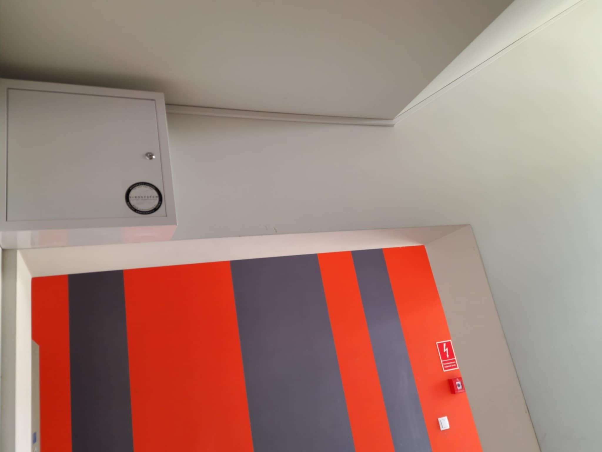 Biała skrzynka z okrągłym logo na ścianie, nad wejściem z pomarańczowo-szarymi pionowymi pasami i symbolem ostrzegawczym z czerwoną błyskawicą.