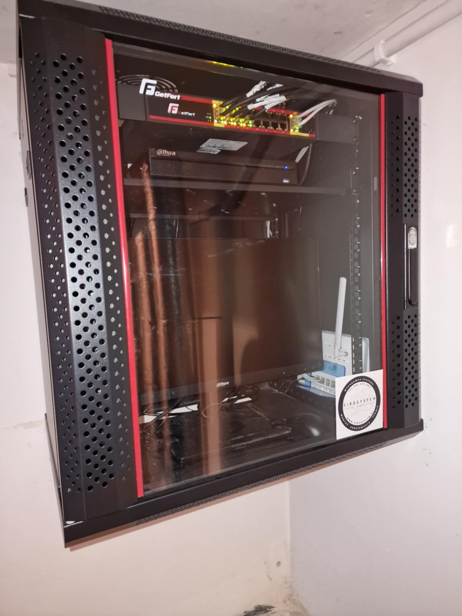 Szafa rackowa z zainstalowanymi urządzeniami sieciowymi GetFort i Dahua, widoczny switch, router oraz biała antena WiFi, na ścianie naklejka z logo firmy SIROSYSTEM.