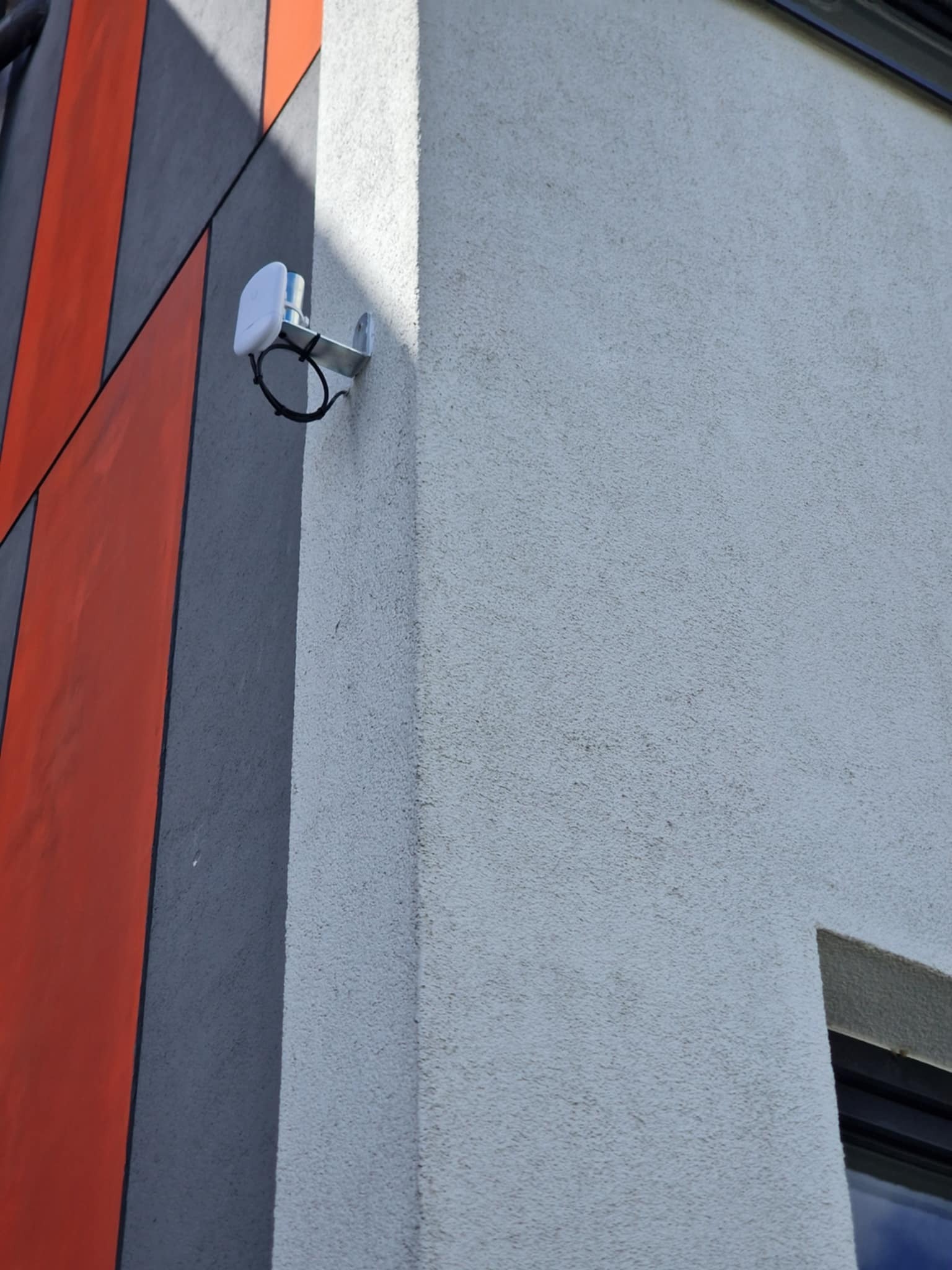 Biała antena Wi-Fi zamontowana na metalowym uchwycie na rogu budynku z szarą i pomarańczową dekoracją.