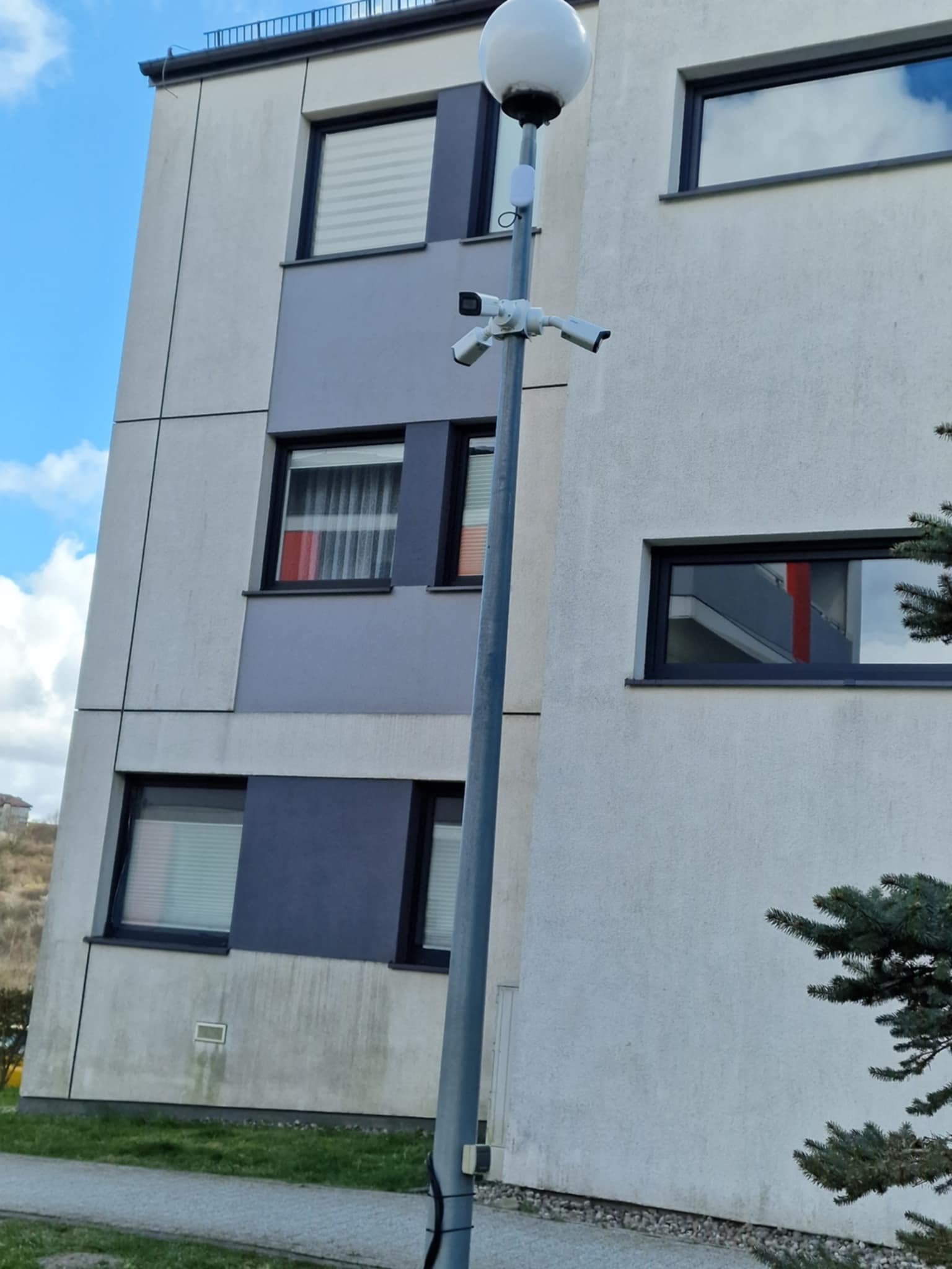 Słup oświetleniowy z trzema kamerami monitoringu na tle budynku wielorodzinnego z widocznymi oknami i fragmentem zieleni.