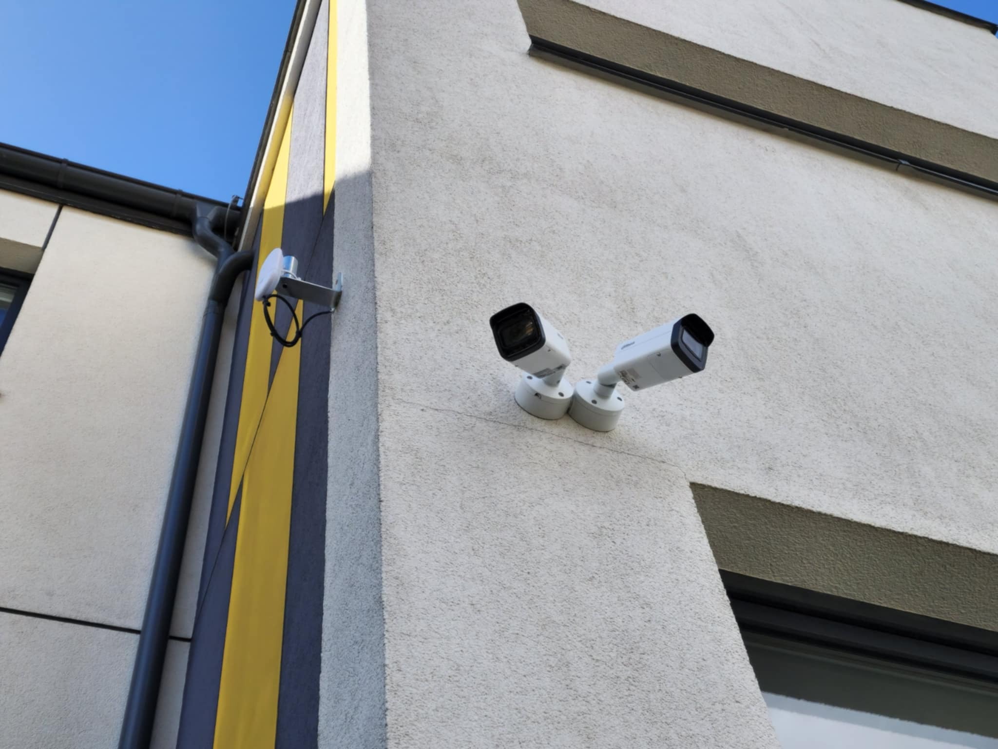 Dwie białe kamery monitoringu na elewacji budynku z żółto-szarym pasem i anteną Wi-Fi, skierowane w przeciwne strony.