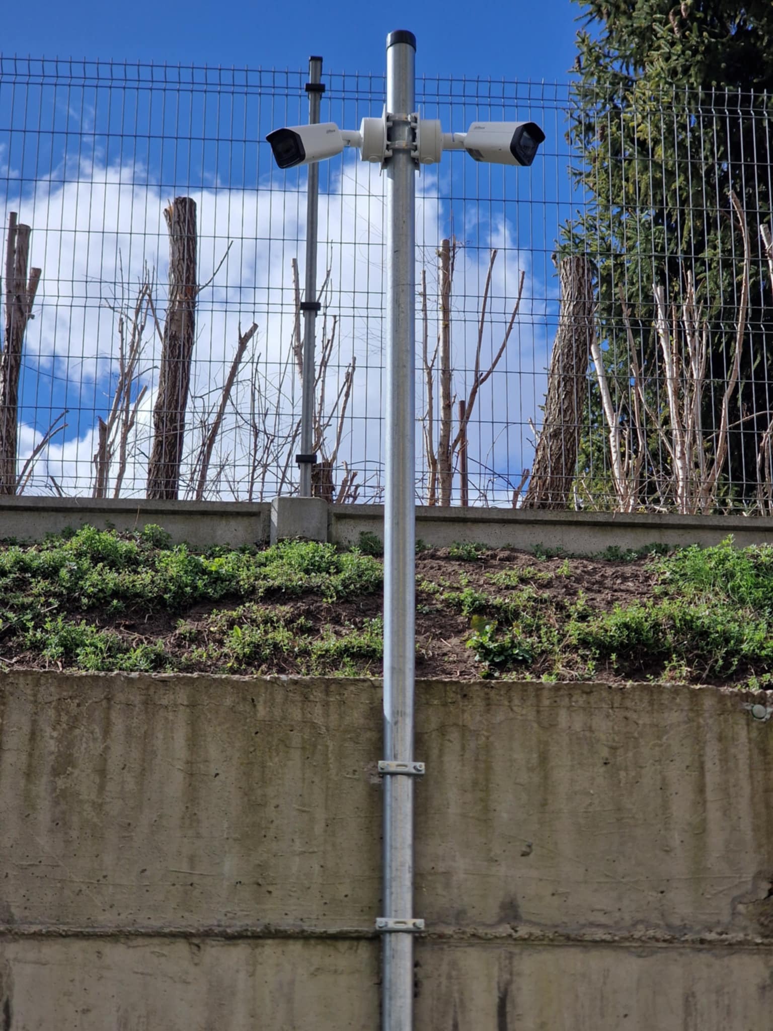 Dwie białe kamery monitoringu zamontowane na metalowym słupie na tle siatki ogrodzeniowej, zieleni i błękitnego nieba z chmurami.