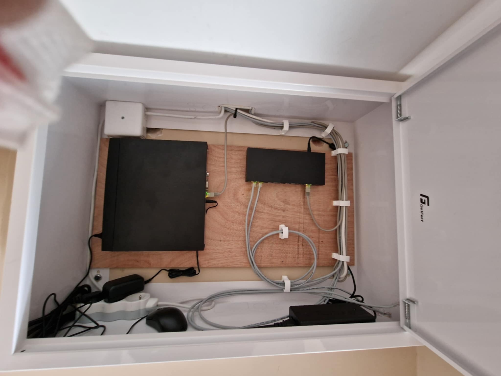 Wnętrze szafki z zamontowanym rejestratorem monitoringu, routerem i okablowaniem, przymocowanym uchwytami do drewnianej płyty, widoczna listwa zasilająca i myszka komputerowa.