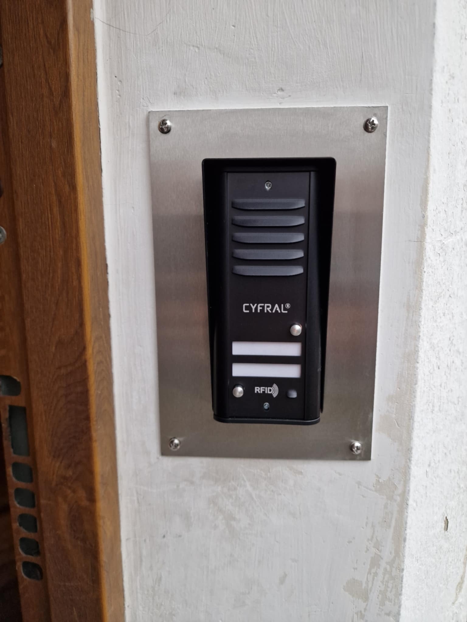 Czarny wideofon marki Cyfral zamontowany na srebrnej, metalowej płycie na pomalowanej, białej ścianie obok drewnianej framugi drzwi.