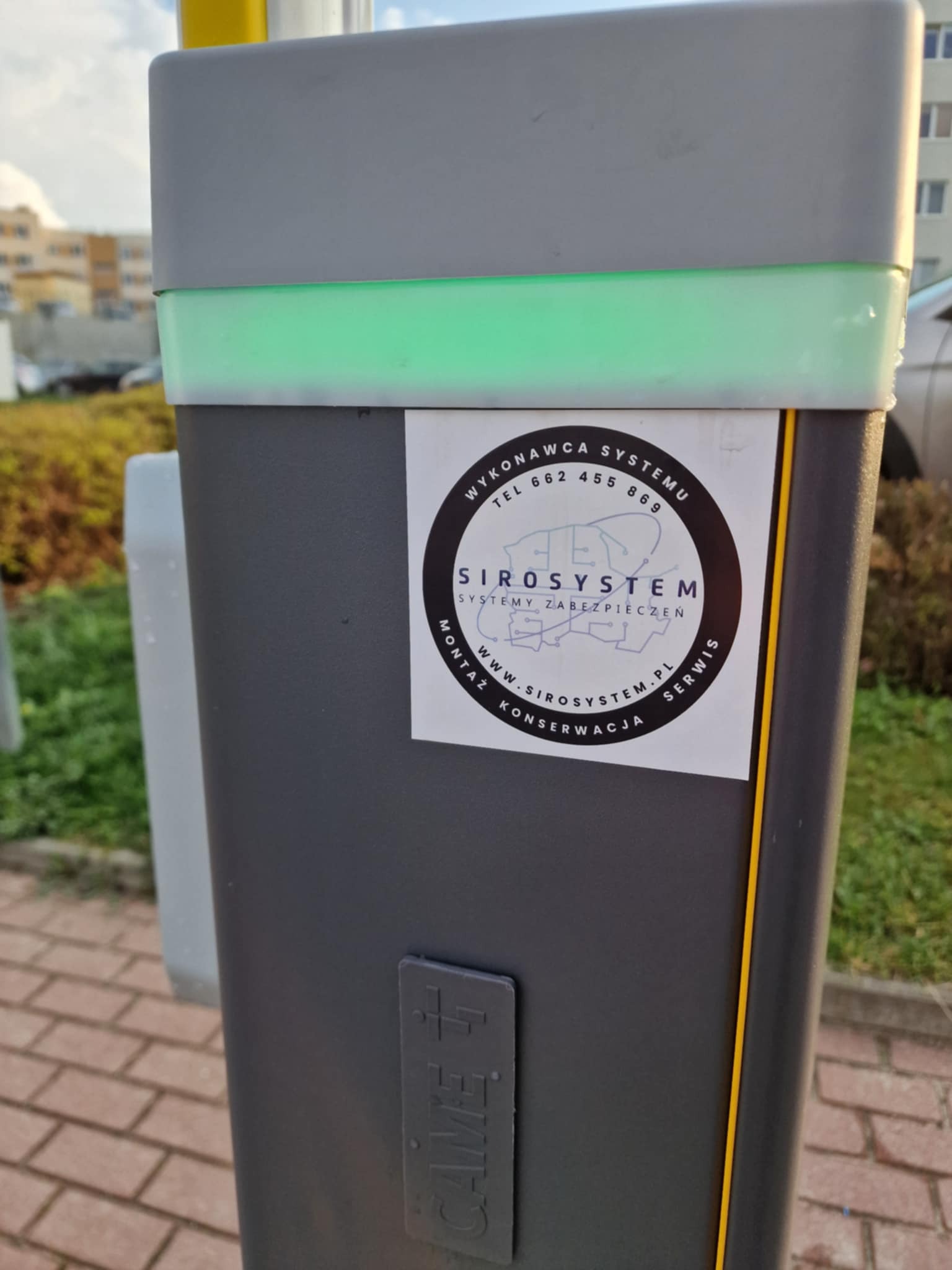 Szary słupek z zielonym podświetleniem i naklejką firmy SIROSYSTEM, zajmującej się systemami zabezpieczeń, widoczny na zewnątrz budynku.