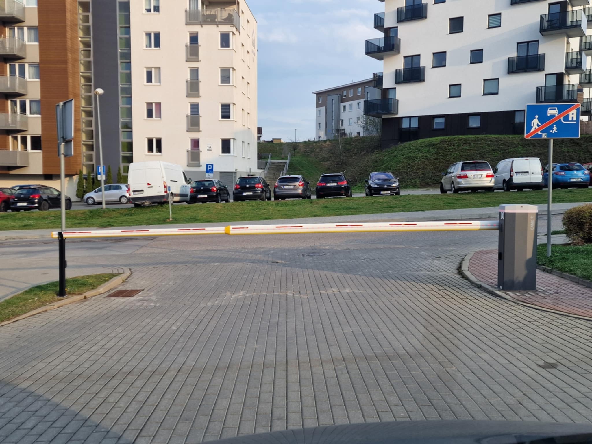 Szlaban automatyczny z żółto-białymi pasami na tle nowoczesnego osiedla mieszkaniowego, zaparkowane samochody, kostka brukowa.