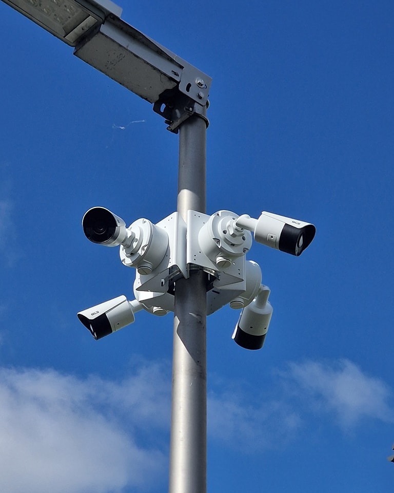 Kompleksowy system monitoringu: cztery kamery CCTV zamontowane na metalowym słupie oświetleniowym, na tle błękitnego nieba z chmurami. Widok z dołu.