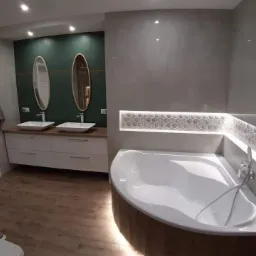 Nowoczesna łazienka z drewnopodobną podłogą, zieloną ścianą z owalnymi lustrami nad białą szafką z umywalkami, oraz narożną wanną z podświetlaną półką wykończoną heksagonalną mozaiką.