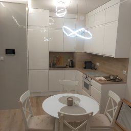 Cezariusz - Nowoczesna, jasna kuchnia z białymi meblami i okrągłym stołem z krzesłami. Uwagę przykuwa designerska lampa sufitowa w kształcie nieskończoności.