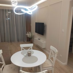 Cezariusz - Jasne wnętrze: biały okrągły stół z krzesłami, minimalistyczny design, telewizor na ścianie z dekoracyjnymi listwami, nowoczesna lampa sufitowa, drewniana podłoga.