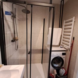 Cezariusz - Nowoczesna kabina prysznicowa z czarnymi profilami w łazience z mozaikową ścianą. Widoczna pralka i grzejnik drabinkowy. Minimalistyczny design.