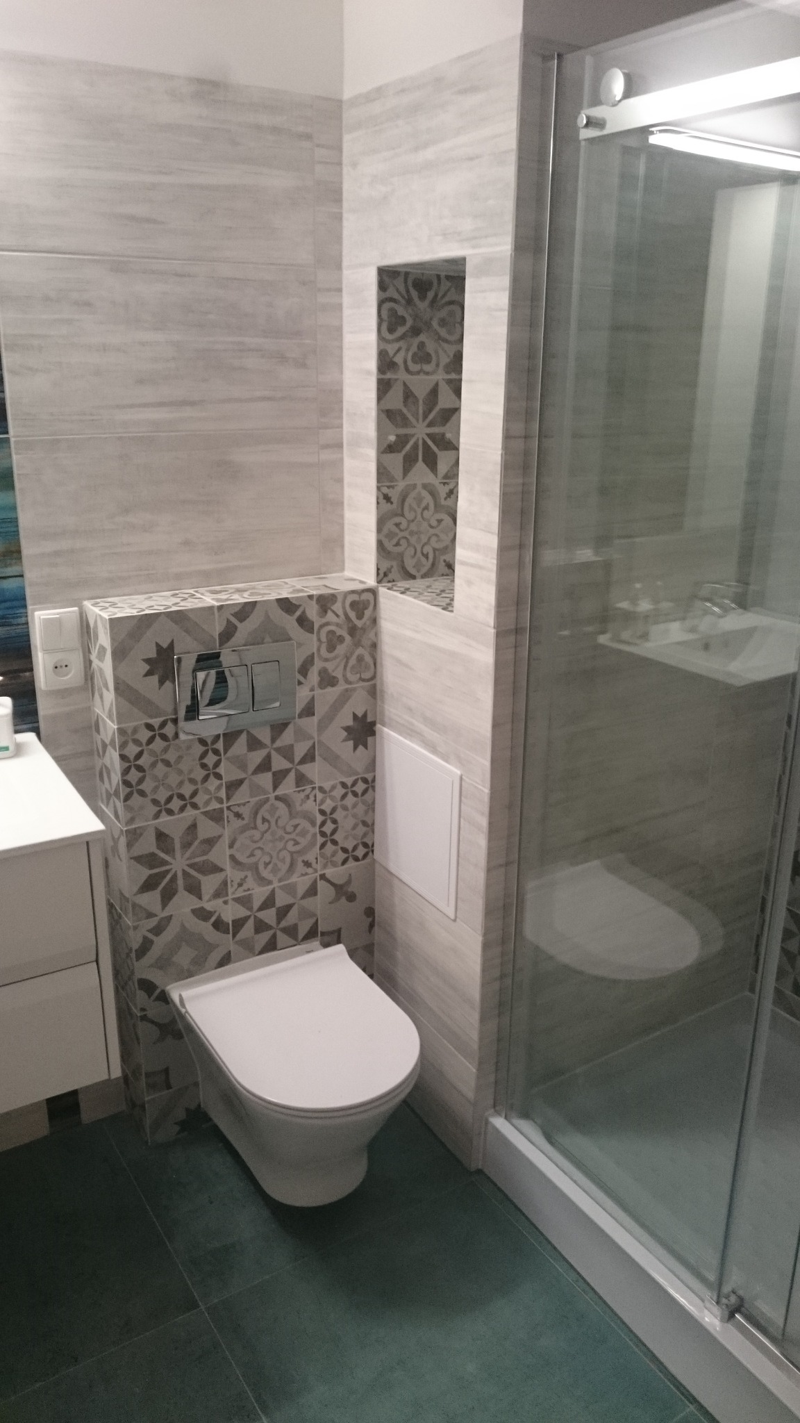 Nowoczesna łazienka z płytkami imitującymi drewno na ścianach, dekoracyjnymi płytkami mozaikowymi przy toalecie i wnęce, oraz kabiną prysznicową ze szklanymi drzwiami.