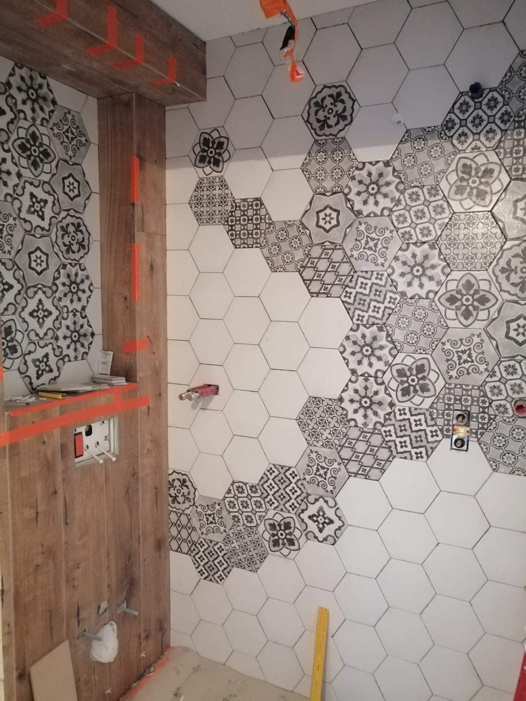 Ściana w trakcie układania heksagonalnych płytek z motywem kwiatowym, część płytek biała, widoczne instalacje hydrauliczne i elektryczne, oraz fragment drewnopodobnej okładziny z pomarańczowymi...
