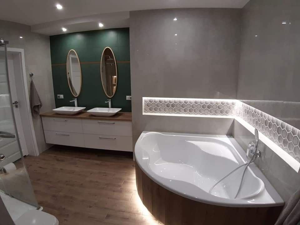 Nowoczesna łazienka z drewnopodobną podłogą, zieloną ścianą z owalnymi lustrami nad białą szafką z umywalkami, oraz narożną wanną z podświetlaną półką wykończoną heksagonalną mozaiką.