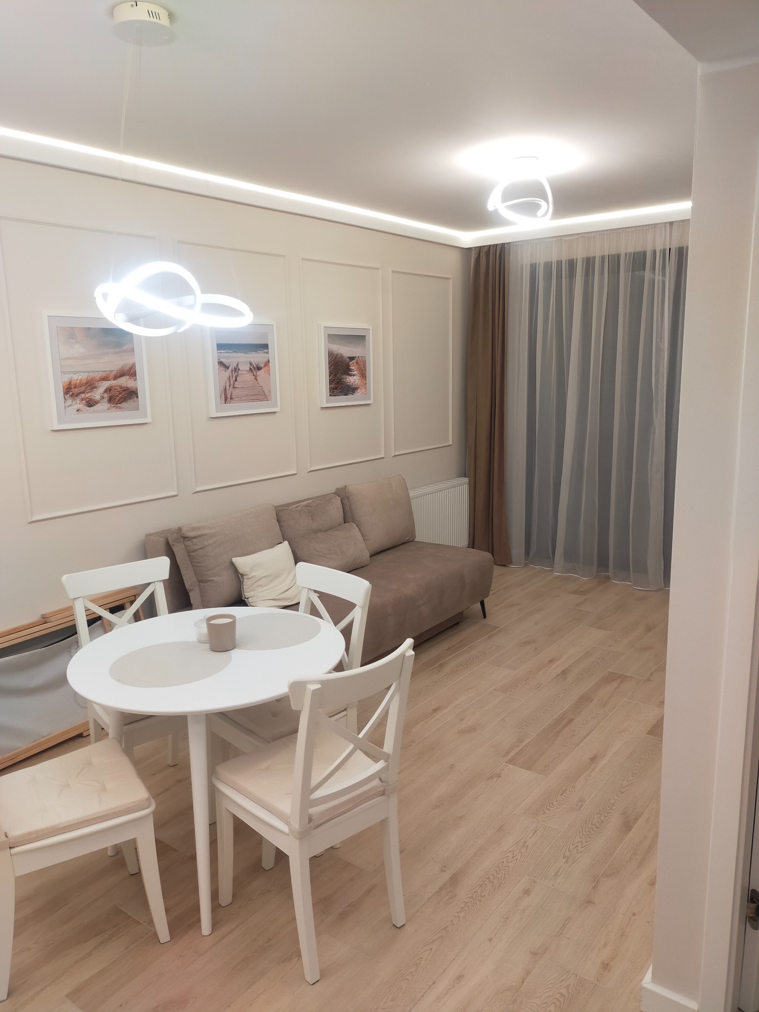 Jasne wnętrze pokoju dziennego z drewnianą podłogą, białym okrągłym stołem i krzesłami. Na ścianie obrazy z motywem plaży i beżowa sofa. Oświetlenie LED.