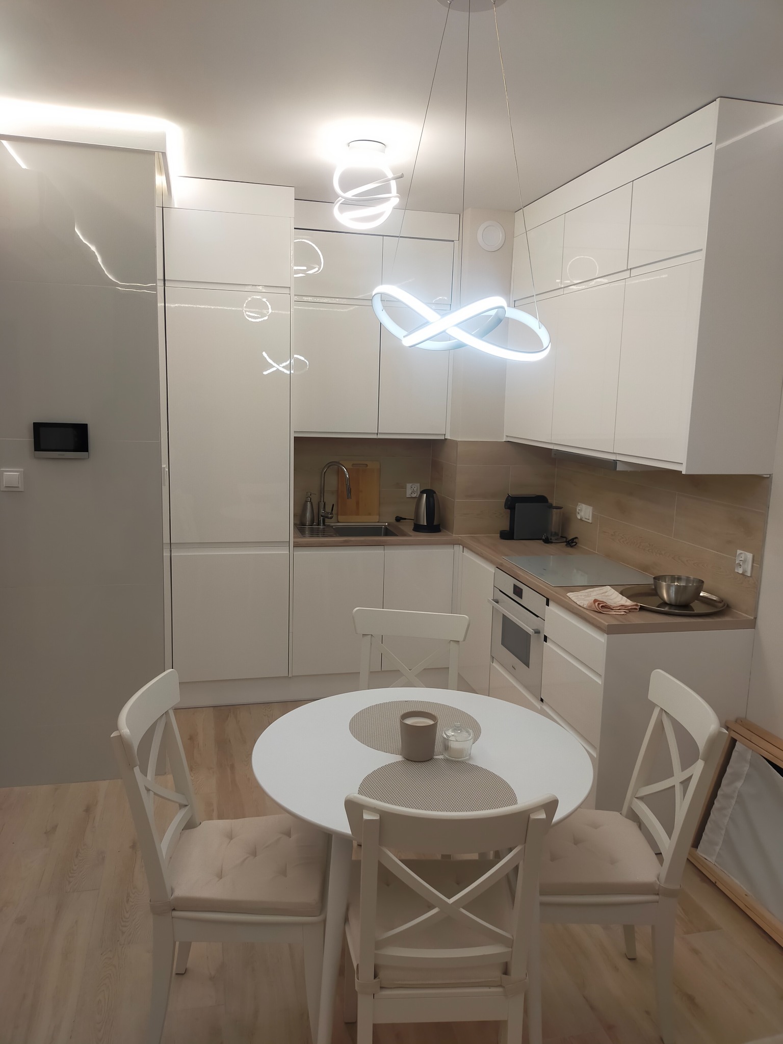 Nowoczesna, jasna kuchnia z białymi meblami i okrągłym stołem z krzesłami. Uwagę przykuwa designerska lampa sufitowa w kształcie nieskończoności.