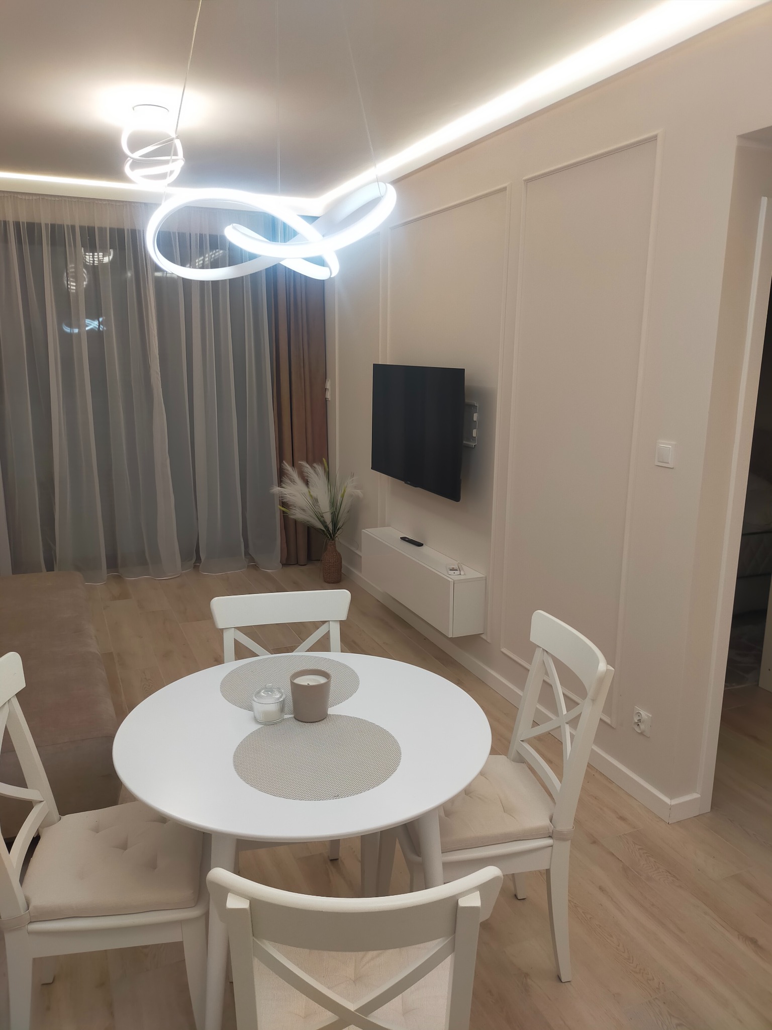 Jasne wnętrze: biały okrągły stół z krzesłami, minimalistyczny design, telewizor na ścianie z dekoracyjnymi listwami, nowoczesna lampa sufitowa, drewniana podłoga.