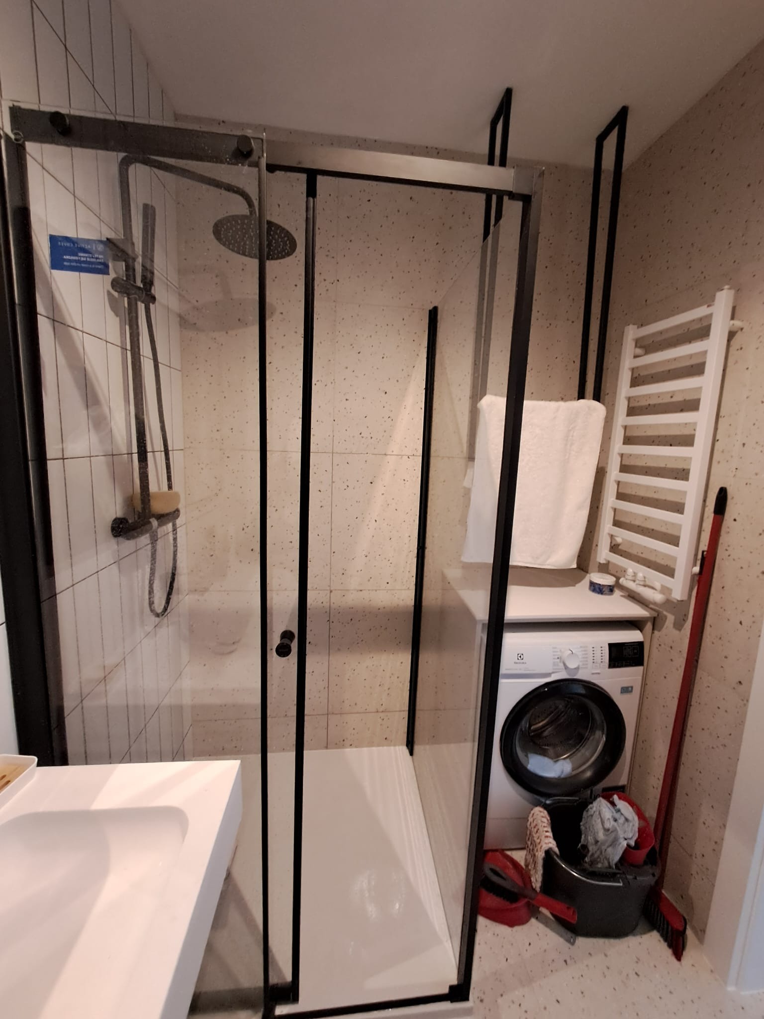 Nowoczesna kabina prysznicowa z czarnymi profilami w łazience z mozaikową ścianą. Widoczna pralka i grzejnik drabinkowy. Minimalistyczny design.