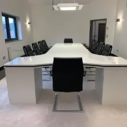 Biały, podświetlany stół konferencyjny o nietypowym kształcie litery T, otoczony czarnymi krzesłami na metalowych nogach, w minimalistycznym wnętrzu z oświetleniem punktowym i wykładziną dywanową.