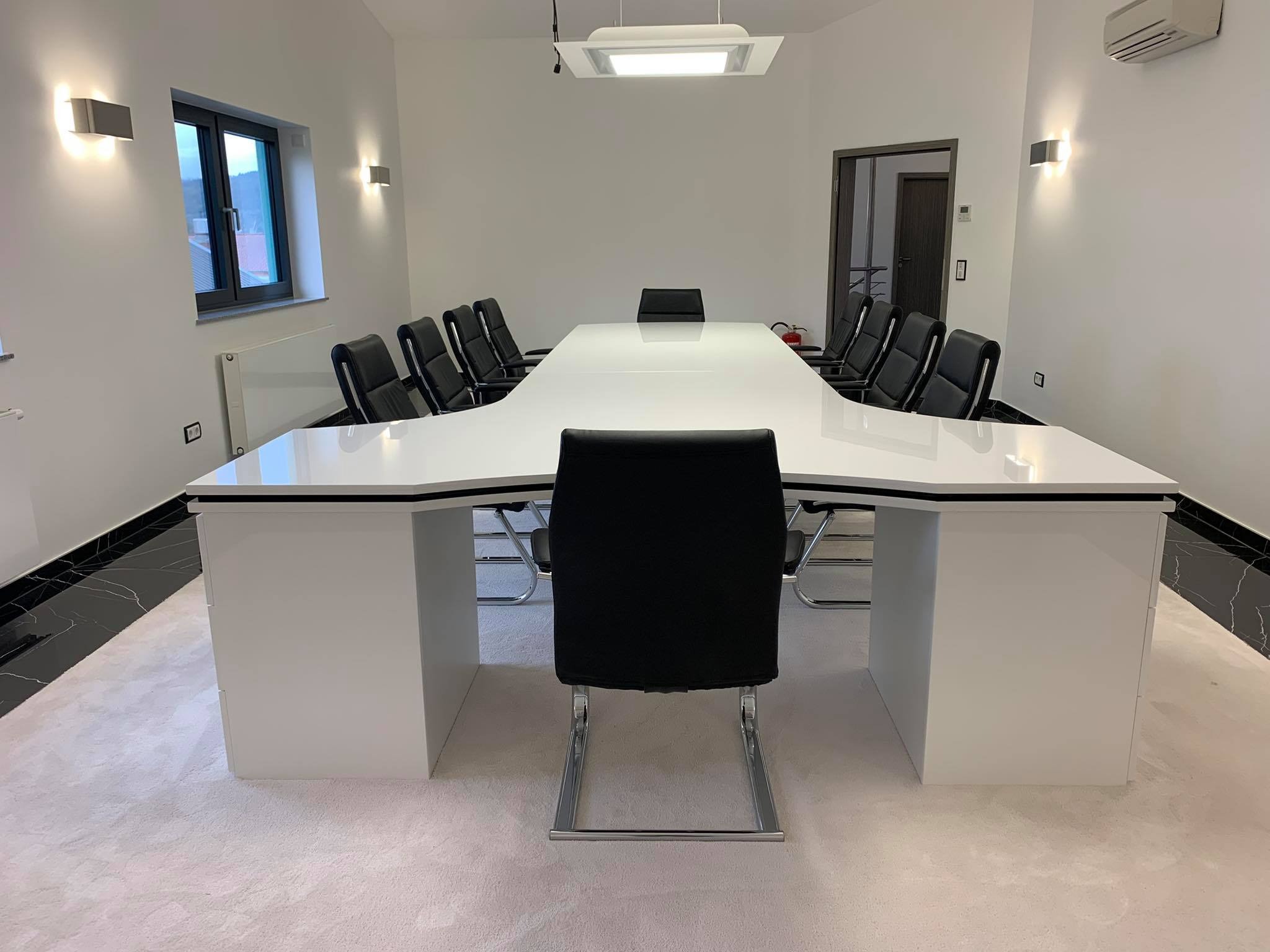 Biały, podświetlany stół konferencyjny o nietypowym kształcie litery T, otoczony czarnymi krzesłami na metalowych nogach, w minimalistycznym wnętrzu z oświetleniem punktowym i wykładziną dywanową.