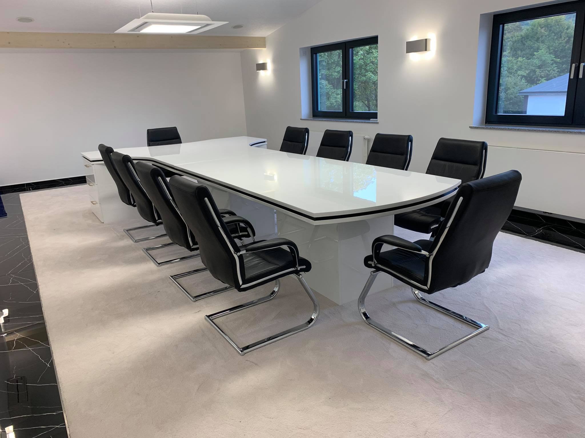 Nowoczesna sala konferencyjna z dużym, białym stołem o zaokrąglonych krawędziach, otoczonym czarnymi skórzanymi krzesłami na chromowanych nogach. W tle widoczne okna z widokiem na zieleń.