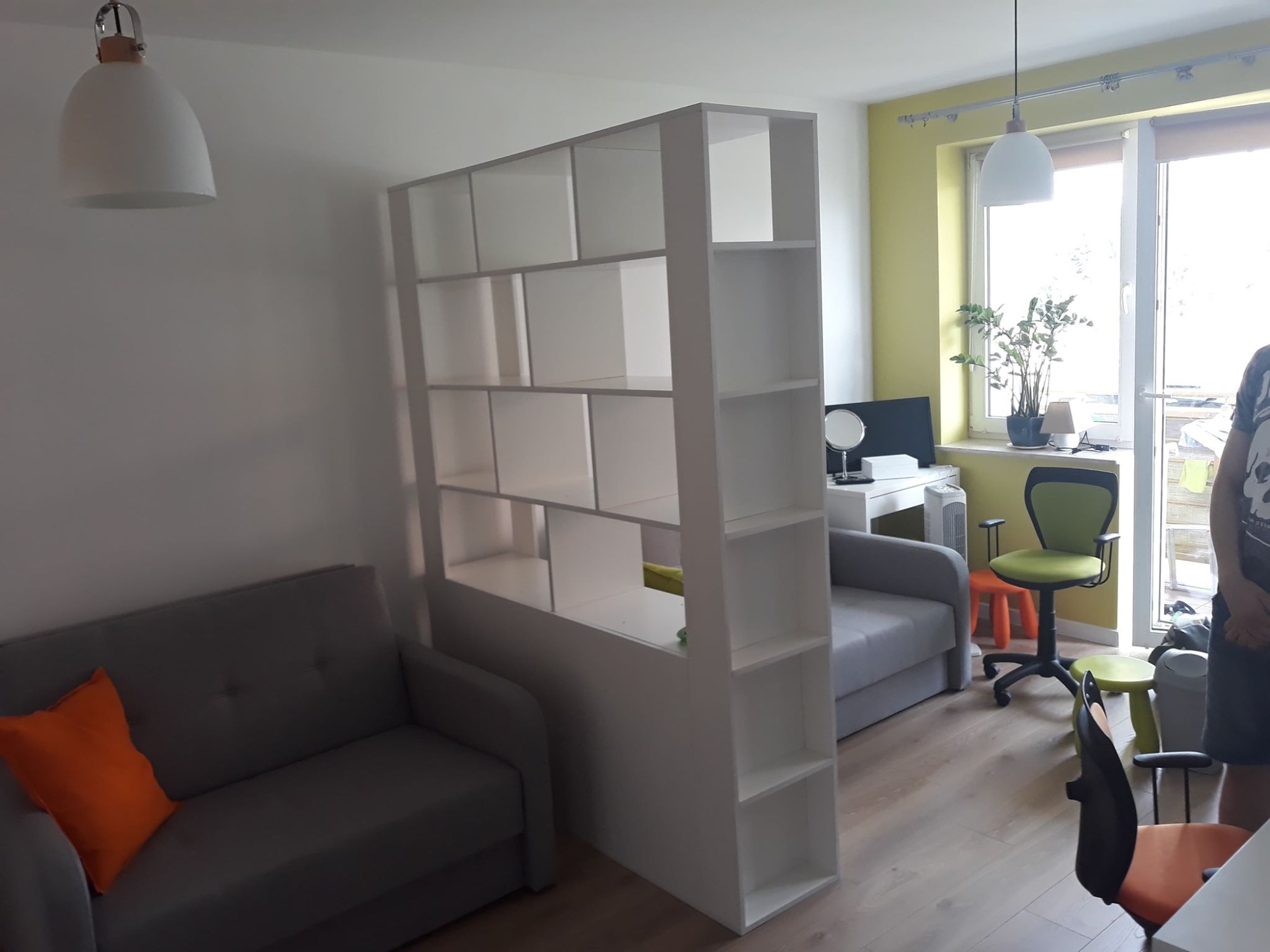 Biały regał z półkami, pełniący funkcję ścianki działowej, oddziela część wypoczynkową z szarą sofą i pomarańczową poduszką od części biurowej z biurkiem i krzesłem. Zwraca uwagę asymetryczny...