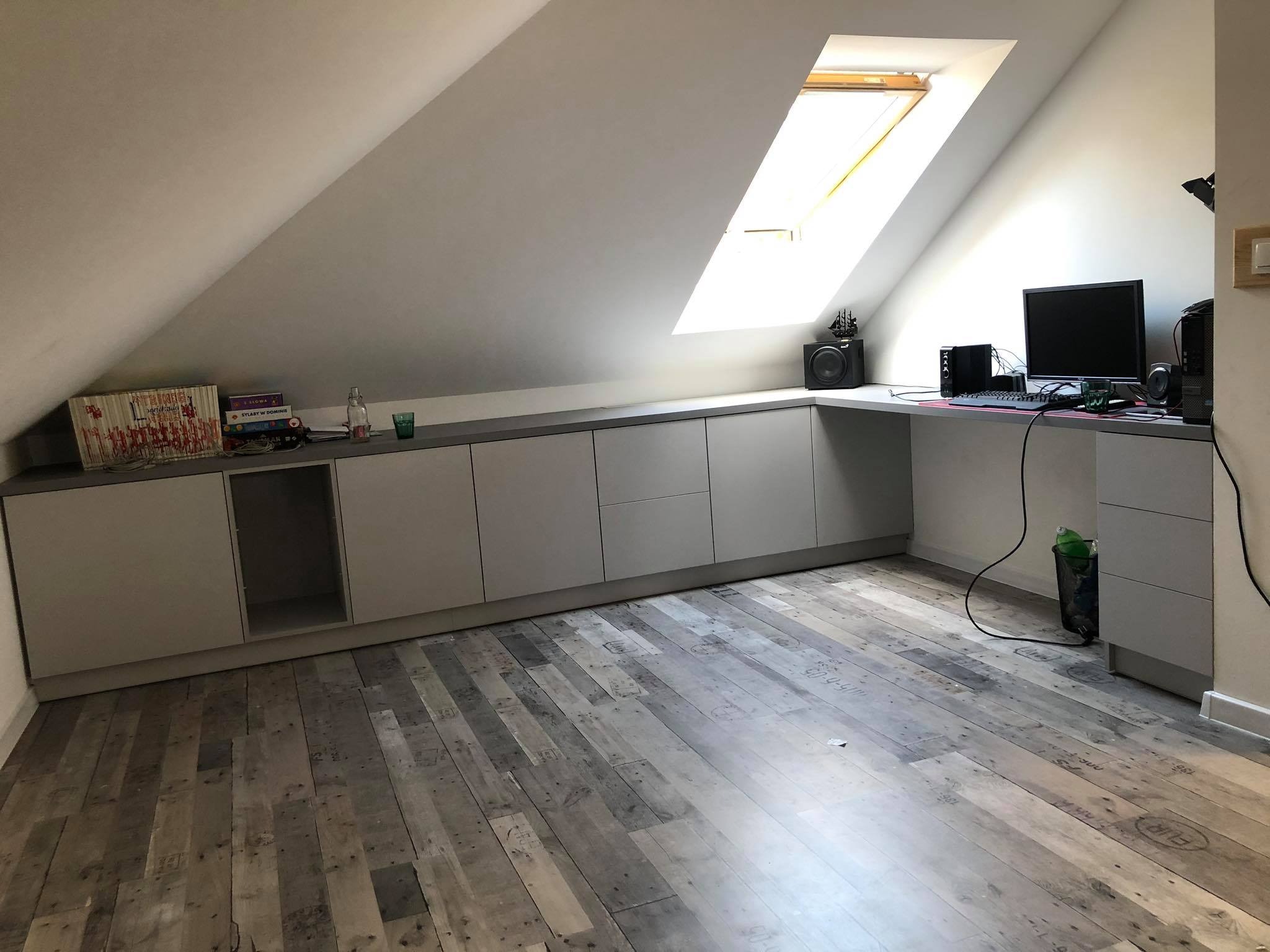 Szara zabudowa meblowa pod skosem na poddaszu z blatem pełniącym funkcję biurka, widoczne książki, głośnik i komputer, podłoga z desek paletowych.