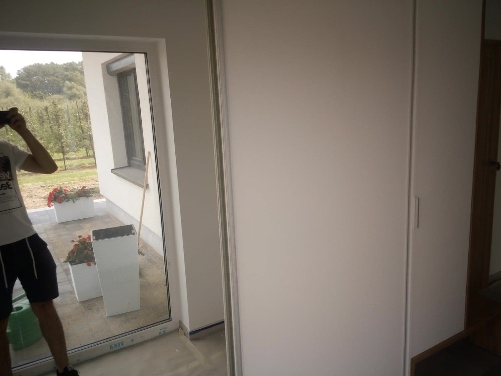 Biała szafa przesuwna z widocznym odbiciem ogrodu przez okno tarasowe. Osoba robiąca zdjęcie widoczna w odbiciu.
