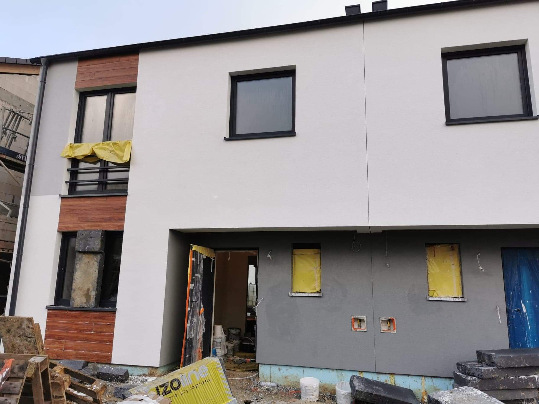 Dwukondygnacyjny dom w trakcie ocieplania elewacji, z częściowo otynkowaną szarą ścianą, zabezpieczonymi oknami żółtą folią i elementami drewnianej okładziny na fasadzie
